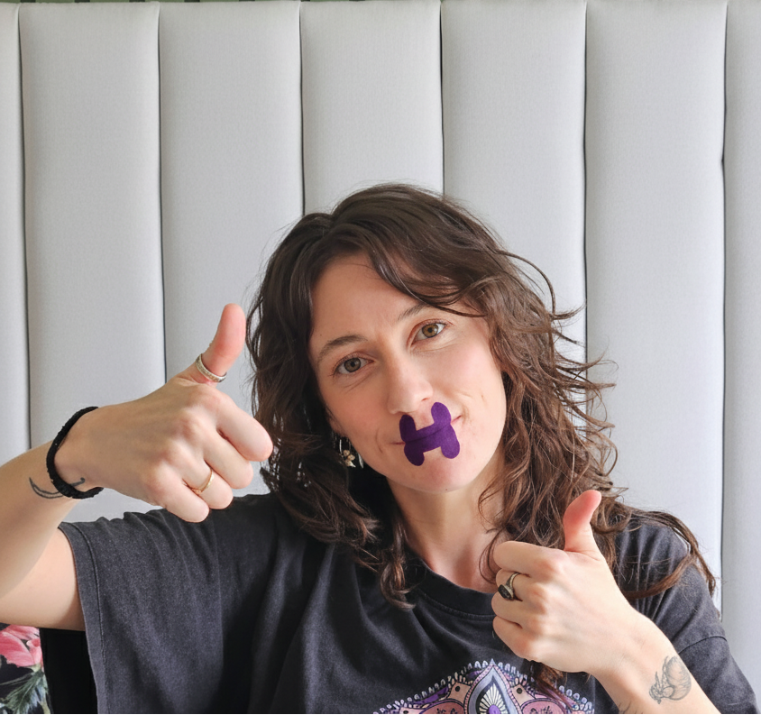 Person with a Hero2 mouth tape on their mouth, giving a thumbs-up gesture in front of a green and gray striped wall.