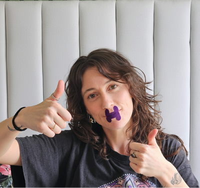 Person with a Hero2 mouth tape on their mouth, giving a thumbs-up gesture in front of a green and gray striped wall.